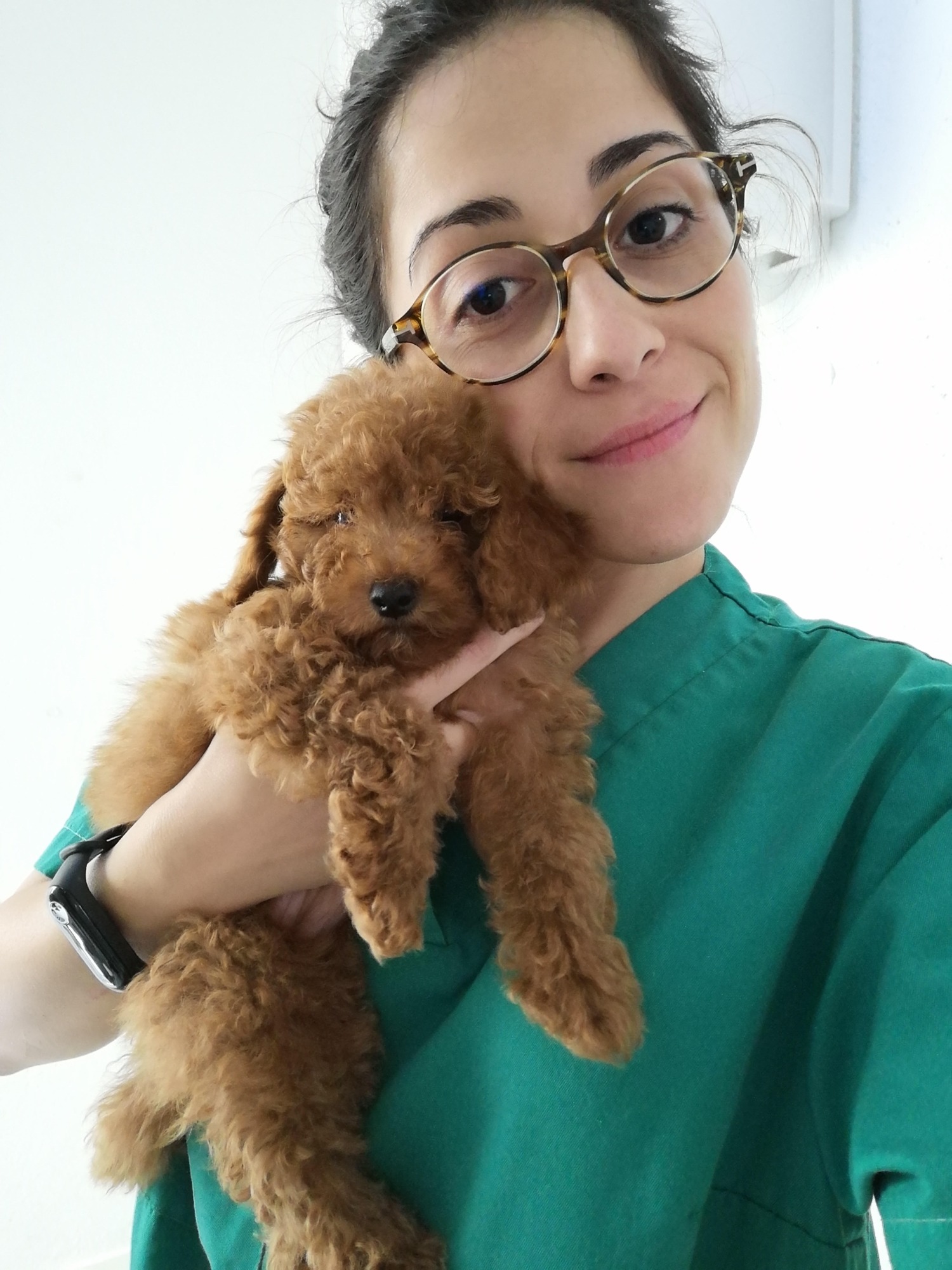 Nuestra veterinaria Mujer con gafas sostiene un cachorro marrón de pelaje rizado, sonriente.